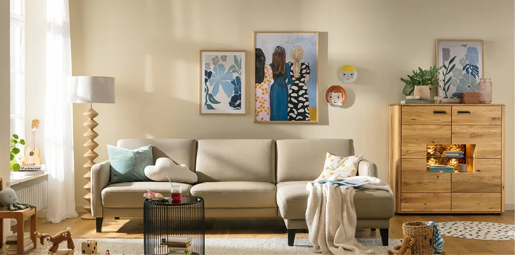 Helles Wohnzimmer mit beigem Ecksofa, Couchtisch und Massivholz-Kommode, dekoriert mit Kissen, Decke und Bildern an der Wand. Gemütliche Wohnidee im modernen Stil mit natürlichen Materialien und warmem Licht.